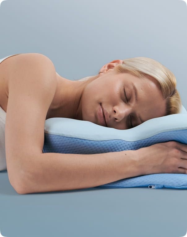 Side sleeper with her arm wrapped around the front of the cervical contour pillow, blue-gray studio background
