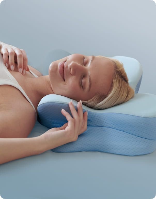 Sleeper resting her head in the contour pillow's centre channel with both hands raised near her shoulder, blue-gray studio background