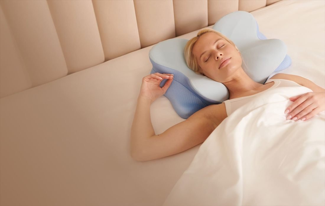 Woman sleeping on her back with her head and neck cradled in the butterfly-contour memory foam pillow, on a bed with a cream upholstered headboard